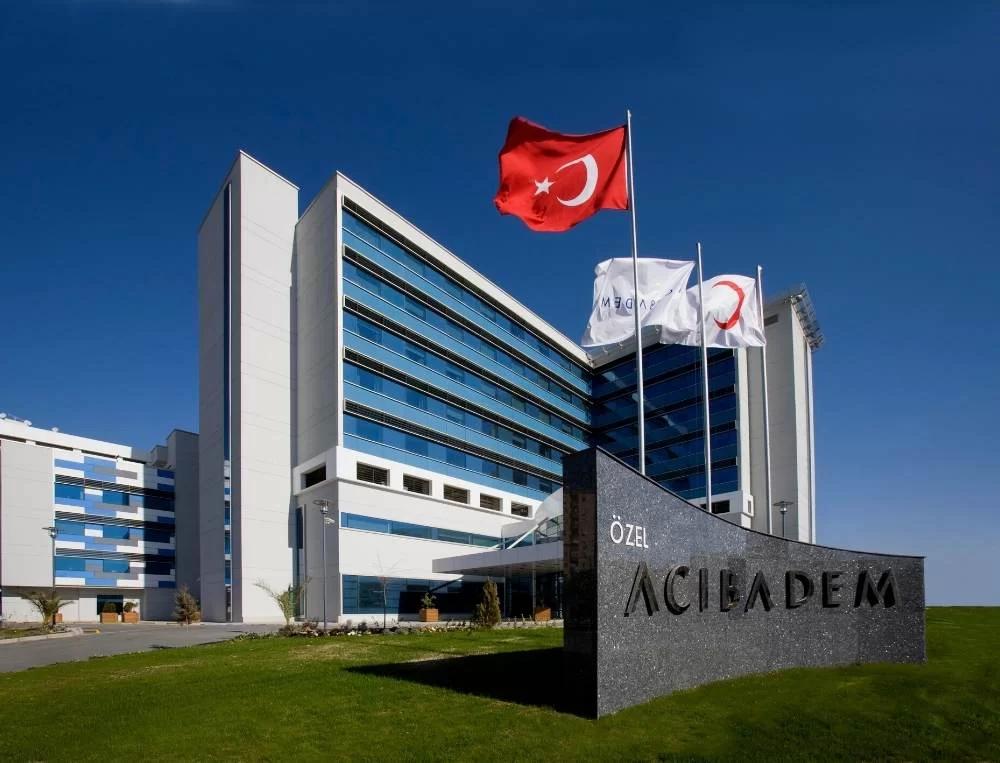 Exterior view of Acibadem Hospital featuring the hospital building, flags, and sign in a green landscape under a clear blue sky.